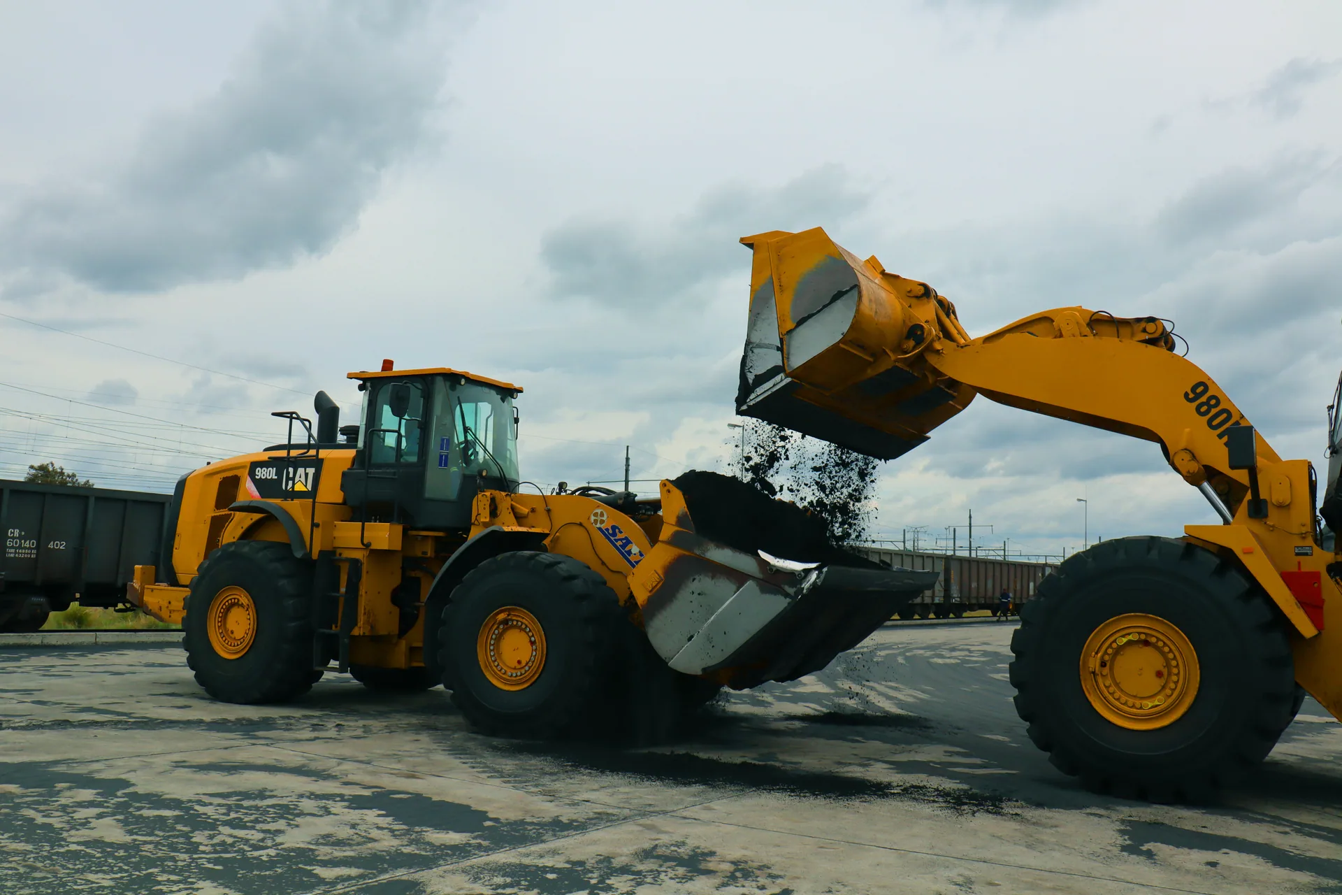 An excavator loading material into the raised bucket of a wheel loader.